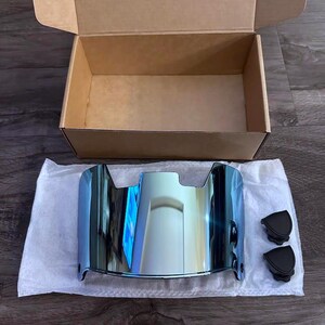 May include: A blue-tinted football helmet visor with a reflective surface, resting on white tissue paper. Two black clips are next to the visor. An open cardboard box is in the background.