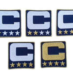 Peut inclure: Écussons brodés avec un "C" blanc sur fond bleu marine, bordure dorée et étoiles dorées. Un écusson a un "C" doré. Les écussons sont de forme carrée.