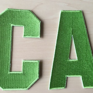 Peut inclure: Deux lettres en feutre, C et A, de couleur vert vif. Les lettres sont cousues ensemble pour former le mot "CA".