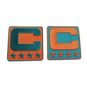 May include: Two embroidered patches. Each patch is a square with a teal border and a teal letter "C" on an orange background. Below the "C" are four teal stars.