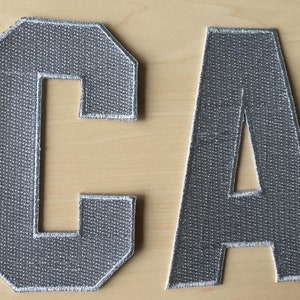 May include: Two gray felt letters, C and A, with white stitching around the edges. The letters are on a light wood surface.