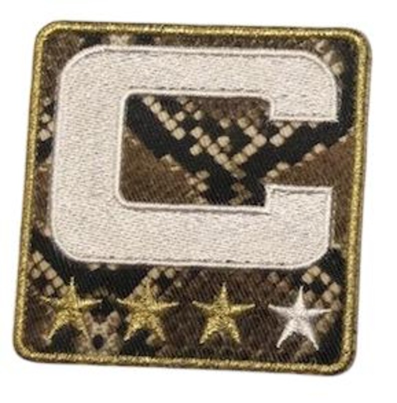 Puede incluir: Parche cuadrado con un fondo de estampado de piel de serpiente, una letra "C" blanca bordada y cuatro estrellas doradas y blancas. El parche tiene un borde dorado.