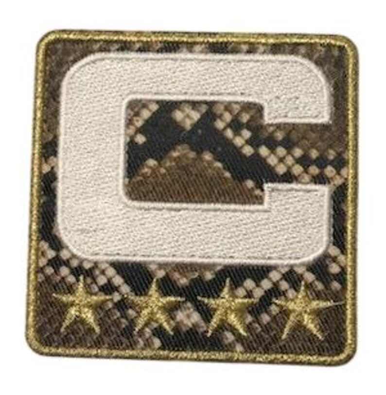 Puede incluir: Parche cuadrado con un fondo de patr&oacute;n de piel de serpiente y una letra "C" blanca bordada. Debajo de la letra hay cuatro adornos dorados en forma de estrella. El parche tiene un borde dorado.