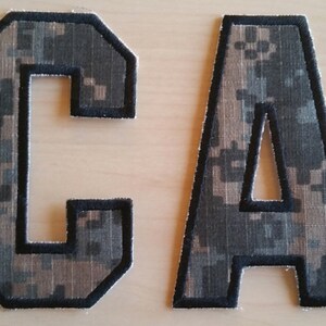 May include: Two large, felt letters, "C" and "A", in a camouflage pattern with black outlines. The letters are sewn together to form the word "CA".