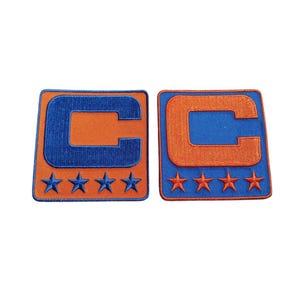 May include: Two embroidered patches featuring a large letter "C" in orange, outlined in blue, on a blue background. Each patch has four stars at the bottom. The patches are rectangular with rounded corners.
