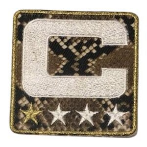 Puede incluir: Parche bordado cuadrado con una gran letra "C" blanca sobre un patr&oacute;n de piel de serpiente. Debajo de la letra hay tres estrellas blancas y una estrella dorada. El parche tiene un borde dorado.