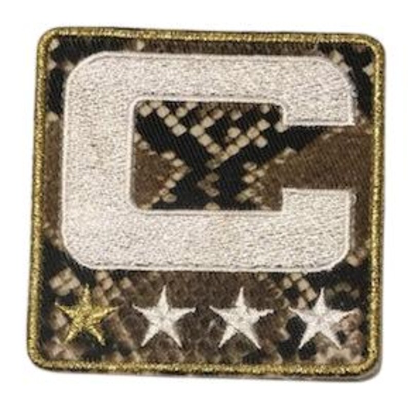 Puede incluir: Parche bordado cuadrado con una gran letra "C" blanca sobre un patr&oacute;n de piel de serpiente. Debajo de la letra hay tres estrellas blancas y una estrella dorada. El parche tiene un borde dorado.