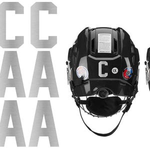 May include: Silver metal letter decals, including the letters "C" and "A", are displayed next to two black hockey helmets. One helmet has a "C" decal, and the other has an "A" decal. The helmets have a chin strap and various stickers.