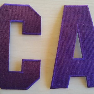 May include: Two purple felt letters, "C" and "A", with white outlines. The letters are sewn together to form the word "CA".