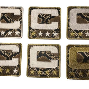 May include: Six embroidered patches featuring a capital letter "C" in various metallic colors, including silver and gold. The patches have a snakeskin pattern background and star accents.
