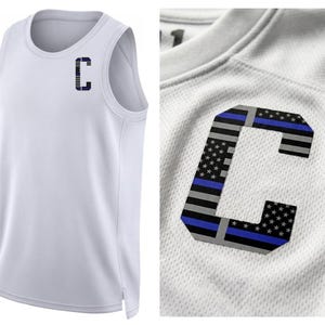May include: White athletic tank top with a large letter "C" on the chest. The "C" features a design incorporating the American flag with blue and gray stripes. The tank top has a round neckline and a sleeveless design.