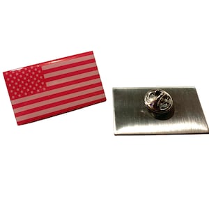 May include: A red, white, and blue American flag pin with a silver backing. The pin is attached to a silver metal backing with a safety pin.