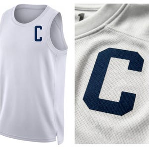 May include: White athletic tank top with a navy blue "C" on the chest. The tank top is made of a breathable, mesh-like fabric. The close-up shows the texture of the fabric and the detail of the letter.