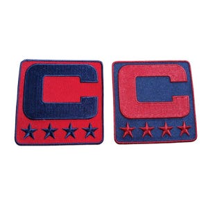 Peut inclure: Deux écussons brodés avec un motif rouge et bleu marine. Chaque écusson présente un "C" rouge sur un fond bleu marine, avec quatre étoiles bleu marine en dessous. Les écussons sont carrés avec des coins arrondis.