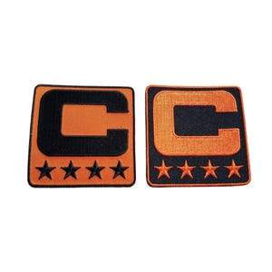 Peut inclure: Deux écussons brodés avec la lettre "C" noire sur fond orange. Les écussons sont carrés avec des bordures orange et quatre étoiles orange en bas. Ces écussons sont probablement pour les vêtements.