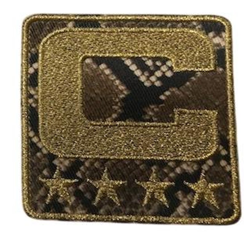 Puede incluir: Parche cuadrado con un fondo de estampado de piel de serpiente y una "C" dorada con purpurina en el centro. Cuatro estrellas doradas con purpurina en la parte inferior. El parche tiene un borde dorado.
