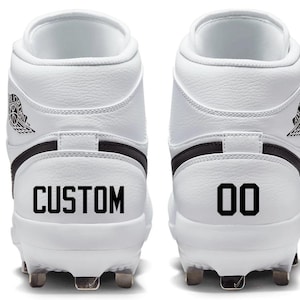 Peut inclure: Paire de chaussures de baseball blanches avec des détails noirs. L'une des chaussures porte le mot "CUSTOM", l'autre le numéro "00". Les chaussures arborent le logo Air Jordan et des crampons métalliques.