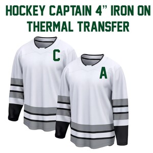 May include: Two white hockey jerseys with black and gray accents. One jersey has a green "C" and the other has a green "A". The text "HOCKEY CAPTAIN 4" IRON ON THERMAL TRANSFER" is at the top.