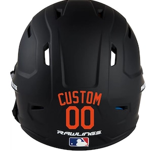 May include: Black baseball helmet with the word "CUSTOM" and the number "00" in orange lettering. The Rawlings logo and MLB logo are also visible. The helmet has several vents and is designed for protection.