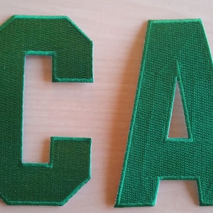 Peut inclure: Deux lettres en feutre vert, "C" et "A", avec un contour blanc. Les lettres sont cousues ensemble pour former le mot "CA".