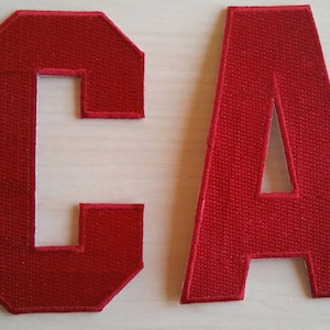 May include: Two red felt letters, C and A, with white outlines. The letters are sewn together to form the word "CA".