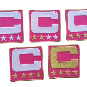May include: Five embroidered patches with a pink background, a white or gold letter "C", and four or five gold stars. The patches have a gold border. The patches are square.