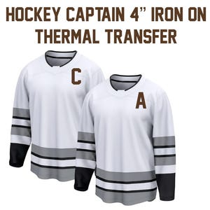 May include: Two white hockey jerseys with black and grey accents. One jersey has a brown "C" and the other has a brown "A". The text "HOCKEY CAPTAIN 4" IRON ON THERMAL TRANSFER" is at the top.