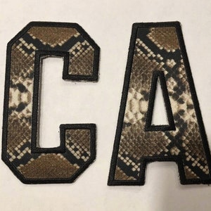 May include: Two felt patches in the shape of the letters "C" and "A". The patches are brown and white with a snake skin pattern and have black outlines.