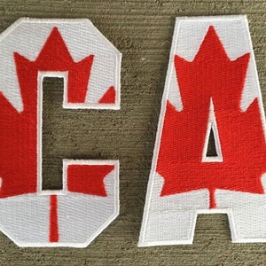 Peut inclure: Deux patchs en feutre blanc avec des motifs de feuilles d'érable rouges, formant les lettres "C" et "A".