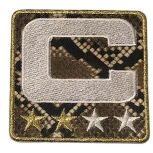 Puede incluir: Parche cuadrado con un fondo de patr&oacute;n de piel de serpiente, con una gran letra "C" bordada en blanco. Debajo de la letra hay tres estrellas bordadas, dos doradas y una plateada. El parche tiene un borde dorado.
