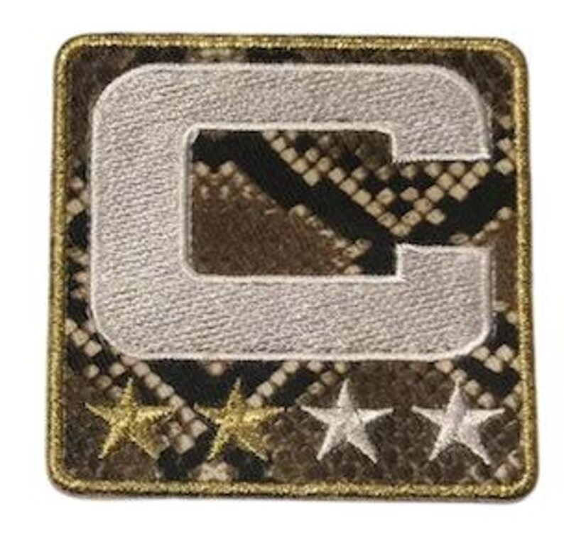 Puede incluir: Parche cuadrado con un fondo de patr&oacute;n de piel de serpiente, con una gran letra "C" bordada en blanco. Debajo de la letra hay tres estrellas bordadas, dos doradas y una plateada. El parche tiene un borde dorado.