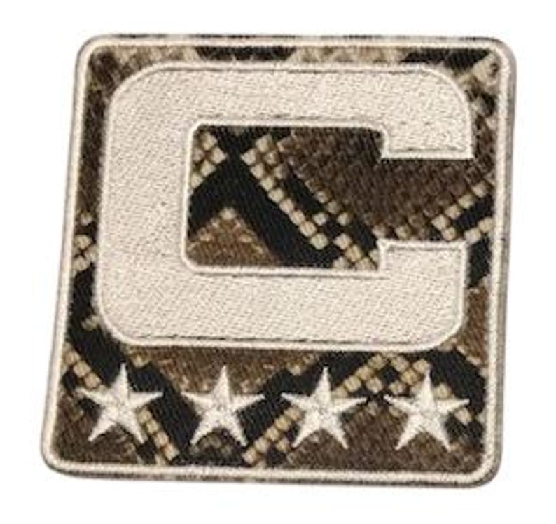 Puede incluir: Parche cuadrado con un fondo de estampado de piel de serpiente y una letra "C" bordada en blanco. Debajo de la letra hay tres estrellas bordadas en blanco. El parche tiene un borde blanco.
