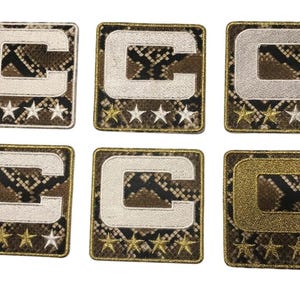 May include: Six embroidered patches featuring a capital letter "C" in various colors: white, silver, and gold. Each patch has a snakeskin pattern background and a row of five stars.