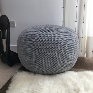 May include: A round, gray crocheted pouf, likely used as a footstool or extra seating. The pouf is made of a textured, knitted material and sits on a white, fluffy rug. The background includes a wall, a curtain, and a dark wooden decorative object.