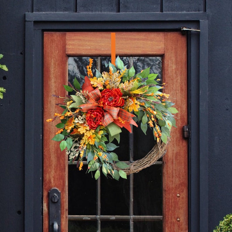 May include: A fall-themed wreath with orange and yellow flowers, green leaves, and a brown woven base. The wreath is hanging on a wooden door with a black frame.