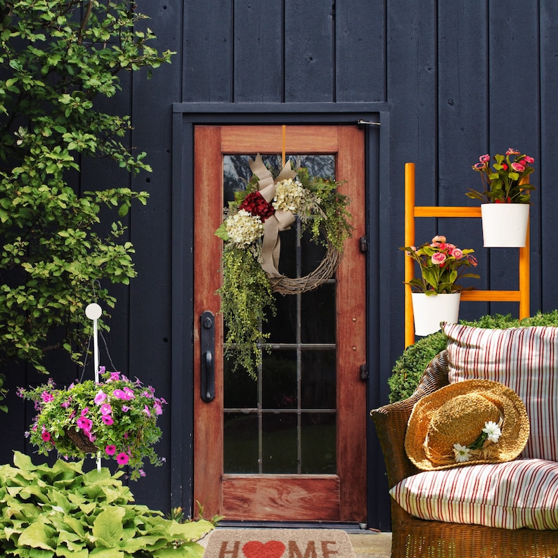 May include: A wooden door with a wreath made of red and white flowers and greenery. The door is painted a dark blue color and has a window with a grid pattern. There is a welcome mat with the word "HOME" on it.