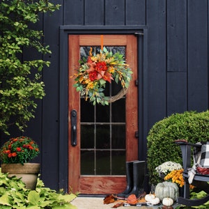 May include: A fall-themed wreath with orange, red, and green leaves and flowers hangs on a dark wooden door. The wreath is made of natural materials and has a rustic look.