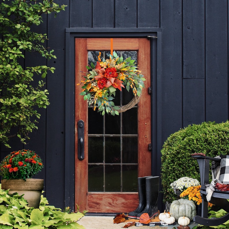 May include: A fall-themed wreath with orange, red, and green leaves and flowers hangs on a dark wooden door. The wreath is made of natural materials and has a rustic look.