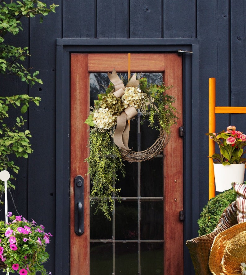 May include: A wreath made of twigs and greenery with white hydrangeas and a brown burlap bow hangs on a wooden door with a glass pane. The door is painted a dark brown color and is set against a dark blue wall.