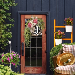 May include: A wooden door with a wreath featuring pink and white flowers and a white letter L. The door is surrounded by a dark blue wall and a wooden ladder with potted plants.