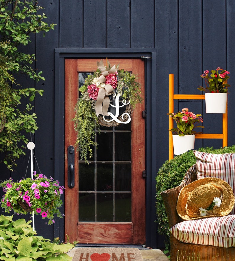 May include: A wooden door with a wreath featuring pink and white flowers and a white letter L. The door is surrounded by a dark blue wall and a wooden ladder with potted plants.