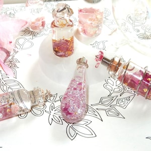 Può includere: Un assortimento di bottiglie e fiale decorative in vetro riempite con accenti rosa e oro. Le bottiglie variano per forma e dimensione, alcune con dettagli in metallo color argento. Il contenuto include petali rosa, glitter e foglie d'oro.