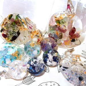 Puede incluir: Una colección de colgantes y esferas de resina transparente, incrustados con piedras preciosas de colores y pan de oro. Los colgantes y esferas varían en tamaño y forma, con una mezcla de azules, verdes, morados y cristales transparentes.