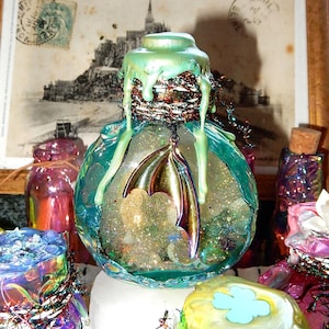 Könnte beinhalten: Eine dekorative Glasflasche mit türkisfarbenem Äußeren und einem grünen, tropfenden Deckel. Im Inneren ist die Flasche mit Glitzer und einem goldfarbenen Fledermausflügel-Anhänger gefüllt. Weitere bunte Flaschen befinden sich im Hintergrund.