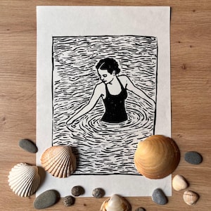 May include: Black and white linocut print of a person in a swimsuit in water. The print is surrounded by seashells and small gray stones on a wooden surface. The person is reaching out with their arms.