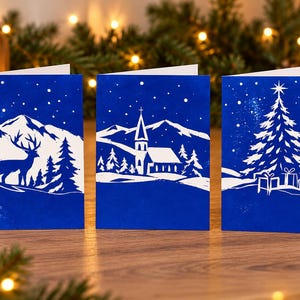 Könnte beinhalten: Drei gefaltete Grußkarten mit tiefblauem Hintergrund und weißen Silhouetten. Die erste zeigt einen Hirsch in einem verschneiten Wald, die zweite eine Kirche in einer Berglandschaft und die dritte einen Weihnachtsbaum mit Geschenken.