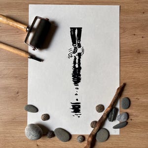 May include: A black ink linocut print on white paper featuring a vertical illustration of legs and feet stepping into a puddle, creating a rippled reflection. The print is displayed with a black rubber brayer, a wooden tool, and several smooth gray stones.