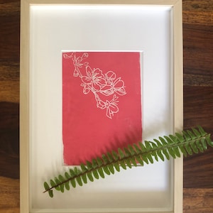 Op de afbeelding: Een ingelijste print van een witte lijntekening van een bloeiende tak op een rode achtergrond. Een groene varen is voor de lijst geplaatst.