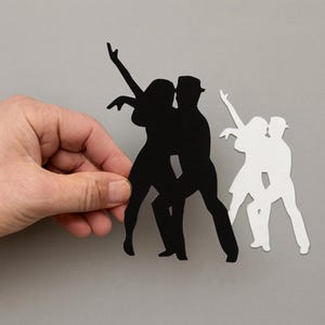 May include: Black and white paper cutouts of dancing couples. One cutout is black, depicting a couple in a tango pose, and the other is white, showing a similar dance position. The cutouts are held by a hand against a gray background.