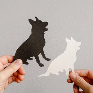 May include: Two paper cutouts of a dog in a sitting position. One is black, and the other is white. The dog silhouettes are held up against a light gray background.